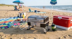 Foldable portable gas grill with collapsible legs set up near beach towels and a cooler